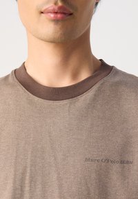 Hnedé tričko s okrúhlym výstrihom vyrobené z mäkkej tkaniny, s kontrastným tmavohnedým ribbed golierom a jemným embosovaným logom.