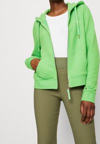 Sudadera verde con cremallera y textura suave, con dos bolsillos frontales y cordones ajustables. Combinada con pantalones ajustados verde oliva.