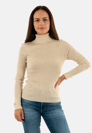 Pullover beige a collo alto a coste con maniche lunghe e dettaglio a bottone sui polsini, abbinato a jeans blu. Texture morbida, design aderente.