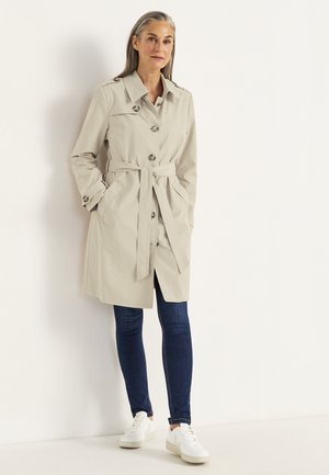 Frau mit grauen Haaren, die einen beigefarbenen Trenchcoat mit Gürtel, dunkelblaue Jeans und weiße Sneakers trägt, stehen vor einer weißen Wand.