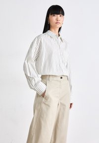 Giovane donna con lunghi capelli neri indossa una camicia bianca con bottoni a strisce sottili nere e pantaloni palazzo beige a vita alta, in piedi contro un muro bianco.