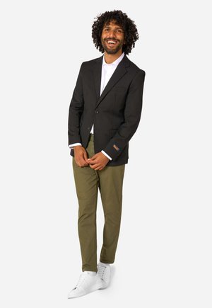 Man met krullend haar die glimlacht, draagt een zwart colbert, een wit overhemd, olijfgroene broek en witte sneakers, staat met gevouwen handen.