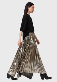 Donna che cammina indossando un top oversize nero, una gonna maxi plissettata metallica e stivaletti neri con tacco contro uno sfondo grigio chiaro.