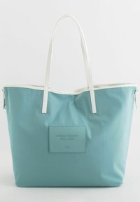 Grand sac fourre-tout teal en tissu résistant avec des poignées blanches et une pièce texturée comportant un texte en relief. Fermeture éclair en haut.