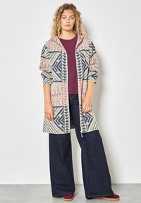 Długi sweter z kapturem, z geometrycznymi wzorami w odcieniach beżu, granatu i różu. Noszony na bordowej bluzce i zestawiony z szerokimi dżinsami.