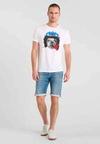 Camiseta blanca con un gráfico de un perro con un casco azul, adornado con estrellas y una rosa. Combinada con unos pantalones cortos de mezclilla y zapatillas blancas.