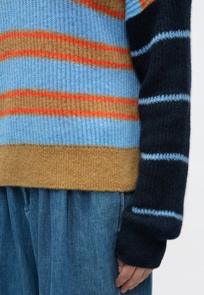 Πολύχρωμο πλεκτό πουλόβερ με ρίγες σε μπλε, πορτοκαλί και καφέ. Μαλακή υφή με φαρδιά εφαρμογή και μακριά μανίκια, συνδυασμένο με τζιν παντελόνι.