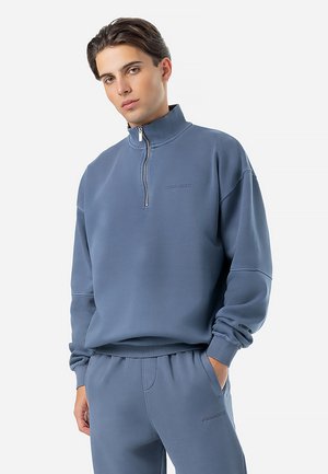 Sweat à capuche zippé bleu clair en tissu doux avec un col montant et un logo brodé. Complété par un pantalon de survêtement assorti, également bleu clair.