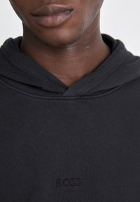 Närbild på en person som bär en svart huvtröja med det broderade ordet "BOSS" centrerat på bröstet.