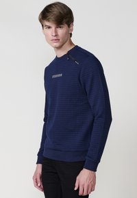 Joven con un suéter azul marino texturizado con pequeños cuadrados y un detalle de cremallera cerca del escote, acompañado de pantalones negros.