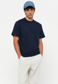 Marineblauwe katoenen t-shirt met een ronde hals en korte mouwen, voorzien van een subtiele logo op de borst. G draagt het met lichtgrijze broeken en een denim pet.