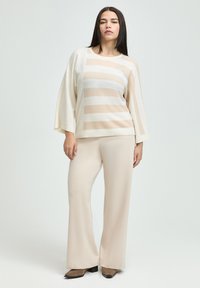 Weicher Strickpullover mit beigen und weißen horizontalen Streifen, im oversized Schnitt mit weiten Ärmeln, kombiniert mit beigen Schlaghosen und braunen Schuhen.