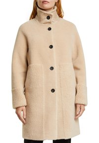 Beige, fluffig kappa med hög krage, sex stora svarta knappar och två framfickor. Designad med ett mjukt, texturerat tyg.