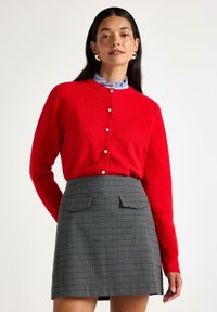 Kvinna iklädd en röd knäppt tröja med pärlknappar, en volangkrage i blått blus och en grå rutig kjol med fickor fram.