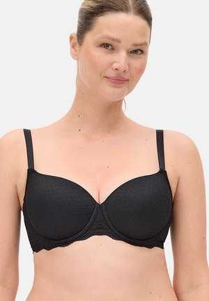 Mujer de piel clara y cabello recogido que lleva un sujetador con copa moldeada negra y ribete de encaje sobre un fondo claro.