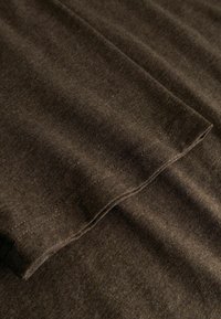 Κοντινό πλάνο καφέ υφάσματος με ορατή ραφή στην διπλωμένη άκρη του μανικιού, που δείχνει τη υφή και τις λεπτομέρειες της ραφής.