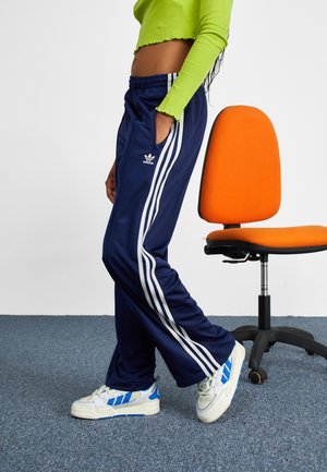 Person iført navyblå Adidas træningsbukser og hvide sneakers med blå striber står ved siden af en orange kontorstol på gråt tæppe.