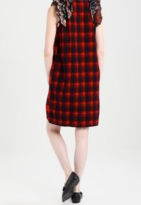 Rotes und schwarzes kariertes Kleid mit lockerer Passform, ausgestattet mit Rüschendetails an den Schultern und einem geraden Saum. Getragen mit schwarzen Slippern.