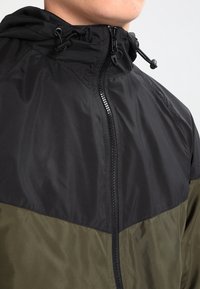 Hooded jacket featuring a black upper section and olive green lower half. Made of lightweight material with a front zip closure and adjustable hood.