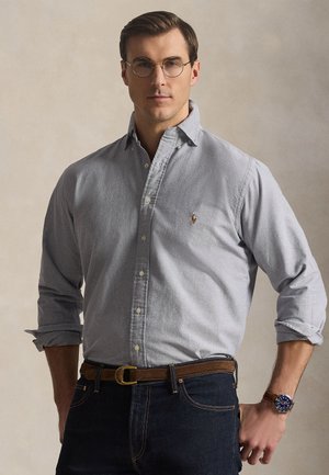 Mężczyzna w jasno szarej koszuli zapinanej na guziki, ciemnych dżinsach, brązowym pasku, okrągłych okularach i niebieskim zegarku na rękę, stojący na tle jednolitego beżowego tła.