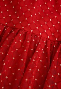 Czerwona tkanina o fakturze z wzorem diamentowym, z elementami złotymi. Tkanina ma falbaniaste krawędzie i jest przezroczysta, lekka w dotyku.