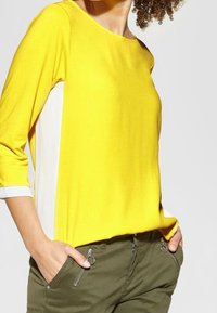 Gul och vit långärmad topp med rund halsringning; mjukt material; avslappnad passform. Gröna byxor med siddragkedjor och fickor.