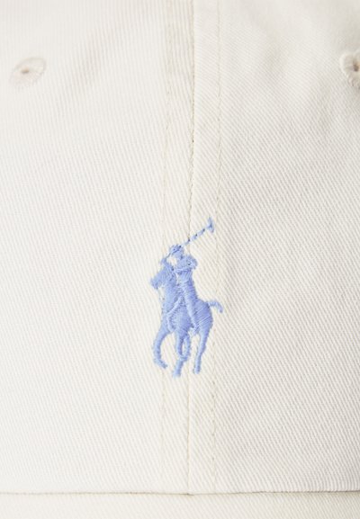 Kremno obarvan material z majhnim modrim vezenim logotipom osebe, ki jaha konja in drži polo palico dvignjeno nad glavo.