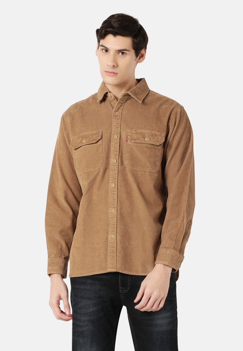 Camisa de pana marrón con cuello abotonado, dos bolsillos en el pecho y una textura acanalada. Combinada con jeans oscuros.