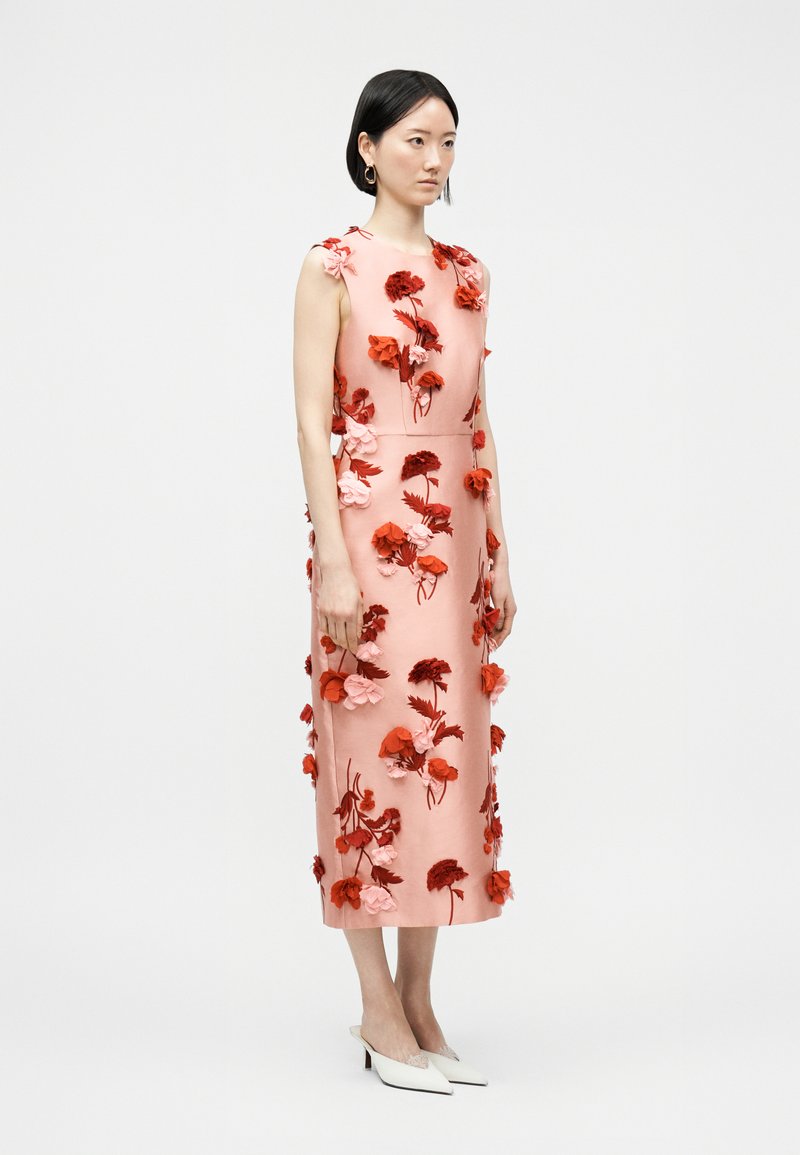 Donna che indossa un vestito midi senza maniche rosa con applicazioni floreali 3D rosse e rosa, abbinato a tacchi bianchi a punta su uno sfondo semplice.