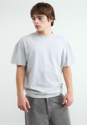 Jeune homme portant un t-shirt oversize gris clair uni et un jean ample gris foncé, regardant vers la gauche sur un fond blanc uni.