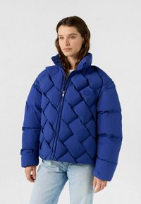 Blaues Puffer Jacket mit diamantgequiltem Design, hohem Kragen und Reißverschluss. Verfügt über ein Logo auf der Brust und übergroße Ärmel.