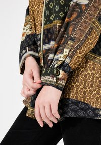 Camisa de patrones multicolores que presenta una mezcla de diseños florales y geométricos, con mangas largas y puños abotonados en tonos de verde, marrón y negro.