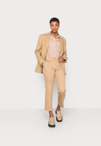 Blazer bege ajustado sobre uma camisa listrada em marrom e branco, combinado com calças cropped bege e sapatos plataforma de cor clara.