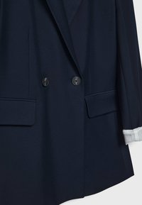 Veste croisée bleu marine à deux boutons noirs, poches à rabat, et un revers de manche laissant apparaître un pull blanc côtelé en dessous.