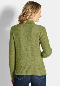 Grüner gestrickter Pullover, strukturiertes Garn mit Sprenkeln, hoher Kragen, gerippter Saum und Bündchen, lockere Passform, kombiniert mit dunkelblauen Jeans.