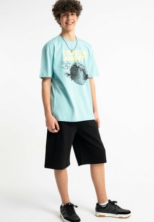 Nastolatek z kręconymi włosami nosi jasnoniebieską koszulkę z grafiką, czarne spodenki do kolan, czarne trampki i łańcuszek na szyi, uśmiecha się i stoi.