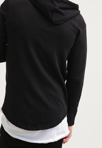 Schwarzer Kapuzenpullover mit langen Ärmeln, ausgestattet mit einem abgerundeten Saum und einem sichtbaren, geschichteten weißen Shirt an der Unterseite. Weicher Stoff.