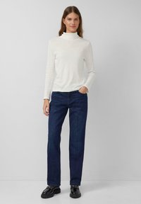 Weißes, eng anliegendes Rollkragenoberteil kombiniert mit dunkelblauen, geraden Jeans. Das Outfit zeichnet sich durch eine glatte Textur und minimalen Hardware aus.