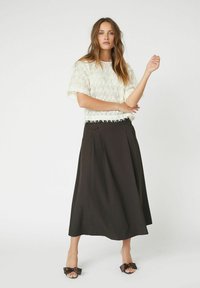 Weißes, strukturiertes Strickoberteil mit Fransenärmeln, kombiniert mit einem langen, schwarzen Rock mit Falten. Das Modell trägt dunkle Sandalen mit Schleifenakzenten.