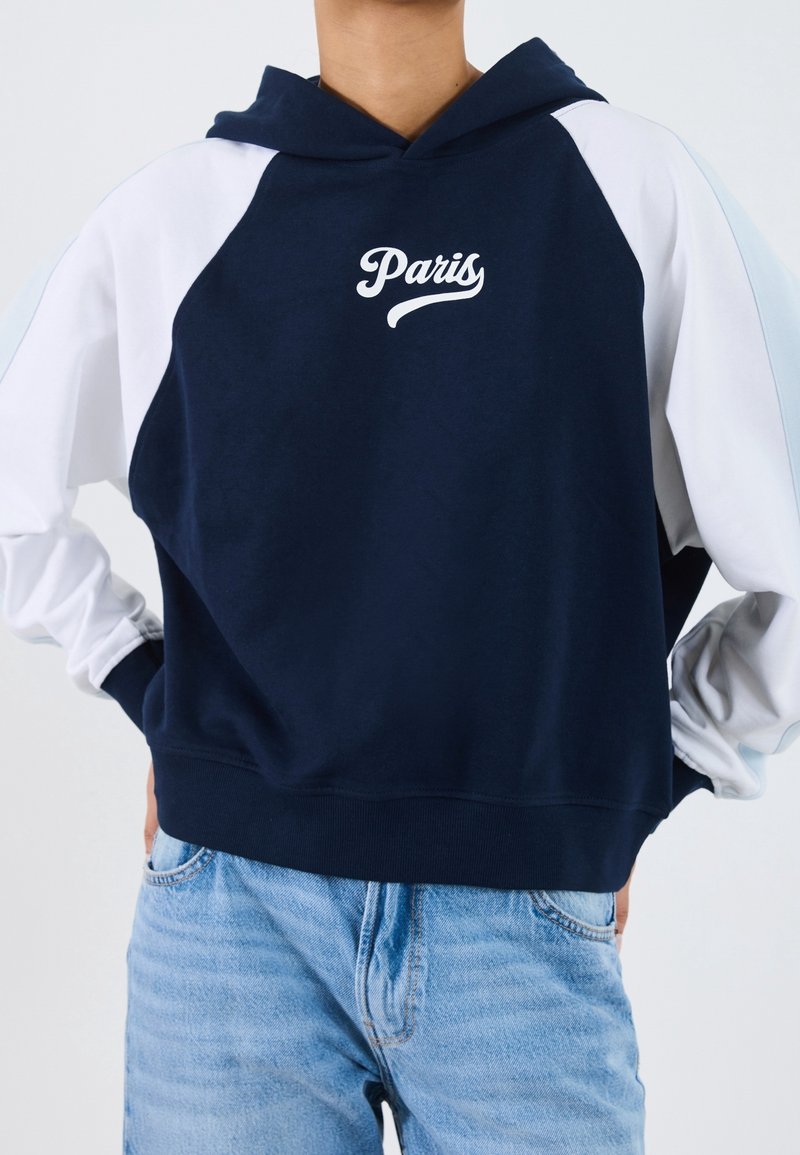 Personne portant un sweat à capuche bleu marine et blanc avec l'inscription "Paris" sur la poitrine, associé à un jean en denim bleu clair, mains sur les hanches.