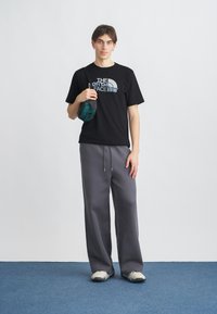 Μαύρο T-shirt με το λογότυπο "The North Face", γκρι φόρμα, και ανοιχτόχρωμα αθλητικά παπούτσια, κρατώντας μια πράσινη θήκη. Ουδέτερο φόντο.