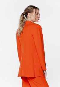 Pomarańczowy dopasowany żakiet o strukturalnym designie, z pojedynczym rozcięciem i slim fit. Posiada gładką teksturę i długie rękawy.