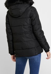 Chaqueta negra acolchada con capucha forrada de piel desmontable, diseño acolchado y detalles de botones a presión en el dobladillo. Mangas largas visibles.