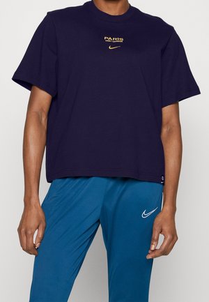 Personne portant un t-shirt bleu marine Paris Saint-Germain et un pantalon bleu Nike montrant les logos sur la poitrine du t-shirt et la cuisse du pantalon.