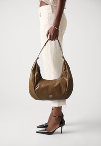 Bolsa hobo de couro castanho com textura brilhante, forma curva e detalhe em logotipo prateado; combinada com jeans brancos cropped e sapatos de salto agulha pretos.