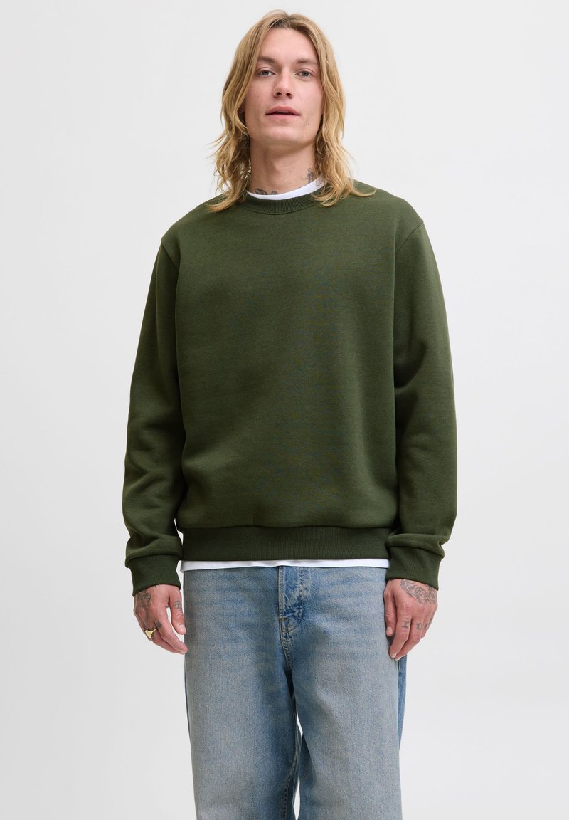 Ung person med axellångt blont hår som bär en olivgrön sweatshirt och ljusblå jeans, stående mot en enkel vit bakgrund.