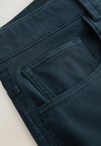 Tamno plavi traper materijal ima glatku teksturu, s malim stražnjim džepom i kontrastnim šavom duž rubova.