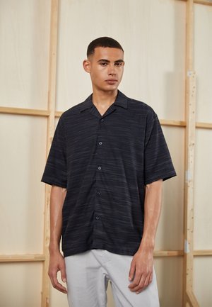 Short-sleeve, dark blue button-up shirt with a textured fabric and horizontal stripe pattern. Displayed on a model with light gray pants.