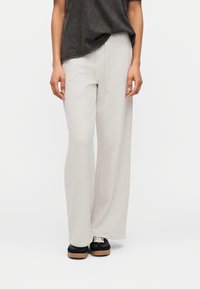 Άτομο που φοράει ανοιχτό γκρι παντελόνια με φαρδιά πόδια και κορδόνι στη μέση, μαύρα και μπεζ αθλητικά παπούτσια, και σκούρο γκρι πουκάμισο.