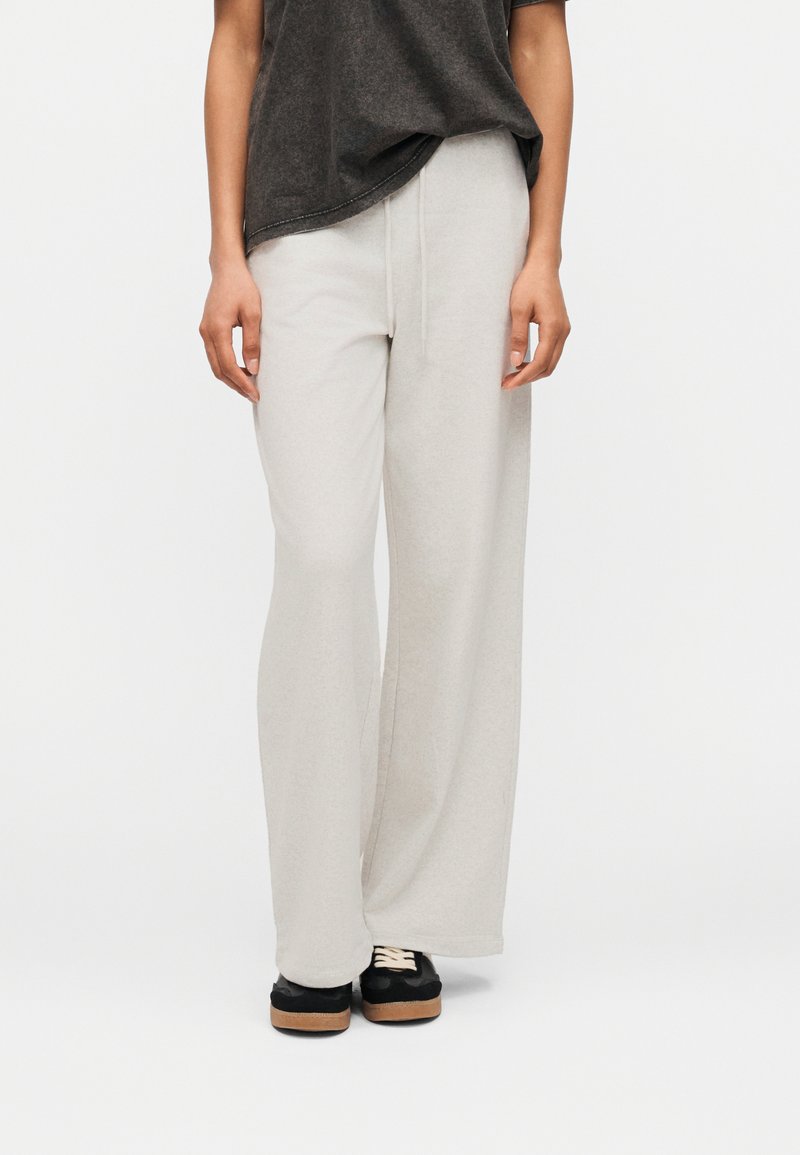 Άτομο που φοράει ανοιχτό γκρι παντελόνια με φαρδιά πόδια και κορδόνι στη μέση, μαύρα και μπεζ αθλητικά παπούτσια, και σκούρο γκρι πουκάμισο.